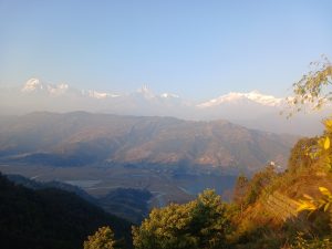 A panoramic view of majestic snow-capped mountains under a clear blue sky, with a misty landscape in the foreground featuring rolling hills, patches of greenery, and a winding river.