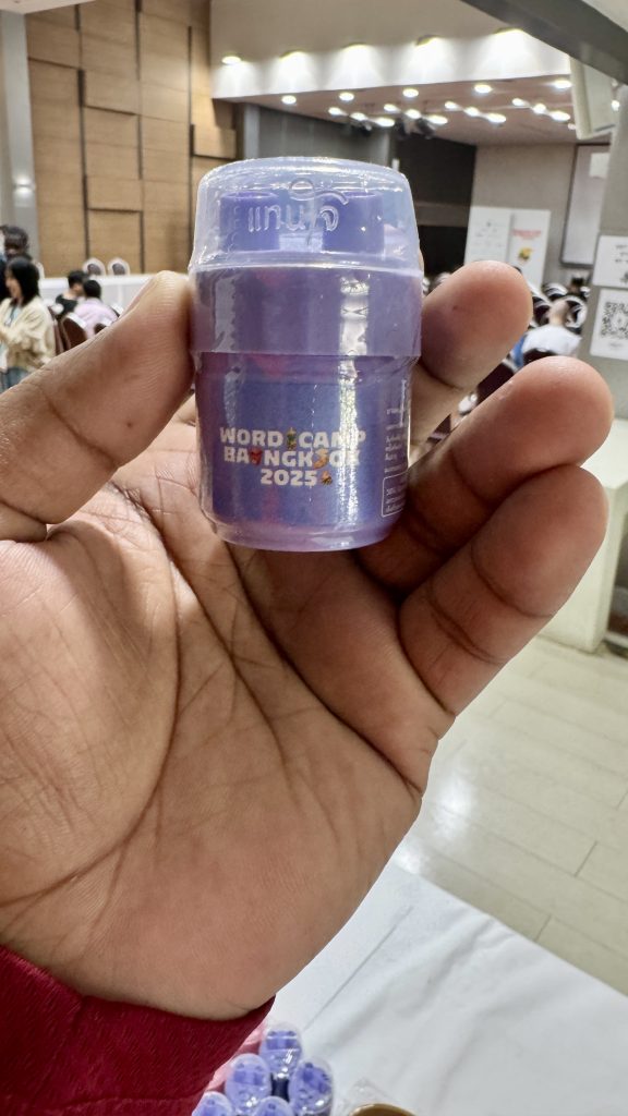 A person’s hand holding a small purple souvenir container or inhaler with the “WordCamp Bangkok 2025” logo printed on the front.
