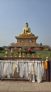A golden Buddha statue sits atop a decorated pedestal, surrounded by offerings such as colourful bowls, flowers, and a small stupa. In the foreground, numerous white scarves known as khada hang from a railing, while a clear blue sky fills the background, creating a serene atmosphere in the open space.