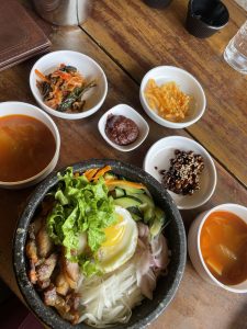 
A stone bowl filled with a variety of ingredients, including sliced vegetables such as cucumbers and carrots, a fried egg with a runny yolk, and pieces of grilled meat.
