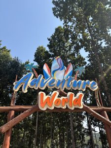 A brightly colored sign over a wooden arch that says "Adventure World". It has stylized palm trees and surfboards around the words.