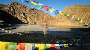 A person stands with arms outstretched beside a frozen lake surrounded by mountains. Colorful prayer flags are strung across the foreground, fluttering in the breeze. 