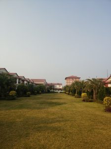 A well-maintained grassy area with neatly trimmed hedges and flowering bushes, flanked by buildings with red roofs. Palm trees and other shrubs are arranged symmetrically along the pathway leading towards a taller building in the background.