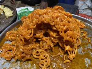 A close-up of bright orange jalebis piled on a metal tray, with syrup glistening to create a warm, indulgent feel.