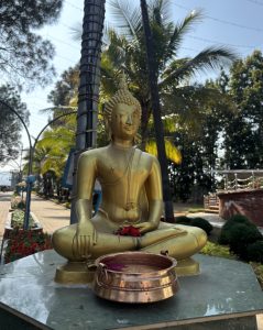 A golden Buddha statue, seated in the lotus position, similar in size to an adult human.  In front is a large copper bowl. In the hands are some red flowers. The setting is a platform set between two sidewalks, there are palm trees in the background.