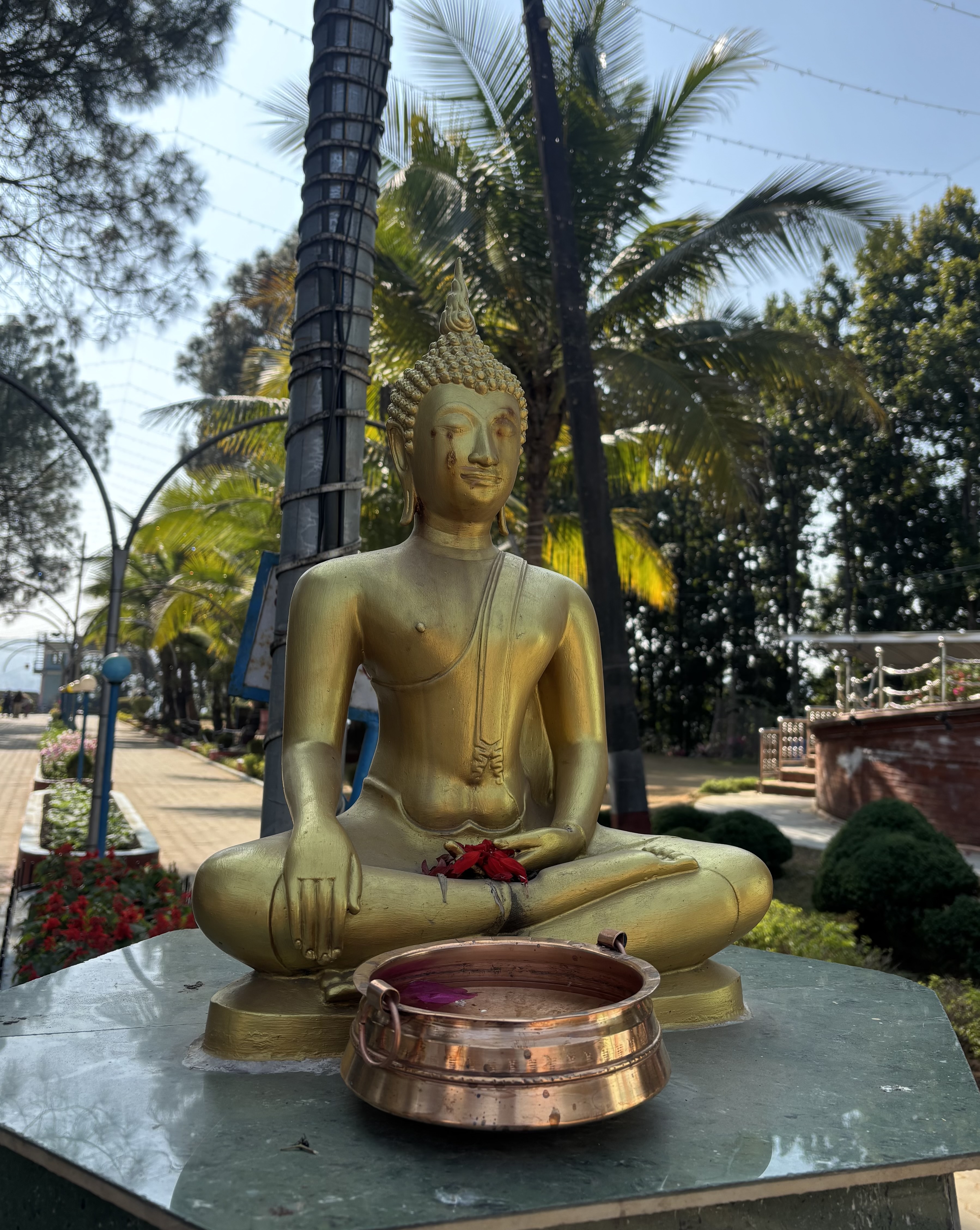 A golden Buddha statue, seated in the lotus position, similar in size to an adult human. In front is a large copper bowl. In the hands are some red flowers. The setting is a platform set between two sidewalks, there are palm trees in the background.