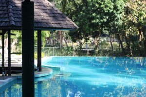 A serene outdoor scene featuring a turquoise swimming pool surrounded by lush greenery. In the foreground, there is a wooden structure with a slanted roof, partially shaded by a supporting column
