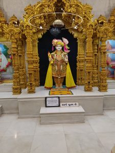 A richly adorned golden temple display at Lord Swaminarayan Nikol Temple features a statue of Swaminarayan in traditional attire, holding a flag in one hand and offering a blessing with the other. The statue is surrounded by intricate golden carvings and illuminated by an elegant chandelier above. In the background, framed portraits of revered figures are displayed, and the floor is finished with polished marble. The serene setting, enhanced by detailed artistry, creates a deeply spiritual and peaceful ambiance.