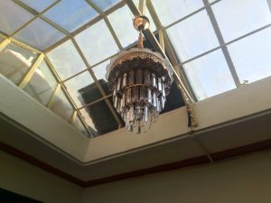 A vintage chandelier hangs beneath a slanted glass skylight with sunlight coming through.