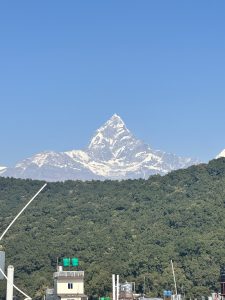 A snow-capped mountain peak rises prominently against a clear blue sky, surrounded by lush green hills.