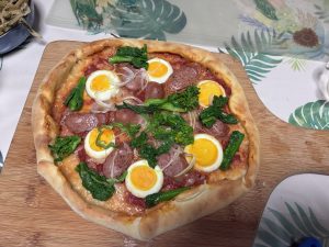 A freshly baked pizza on a wooden cutting board, topped with slices of sausage, halved boiled eggs, and blanched greens. 