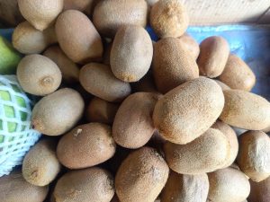 A close-up view of a pile of brown, fuzzy-skinned fruits resembling kiwis, with a few green fruits partially visible to the side, and the overall setting featuring a textured surface.