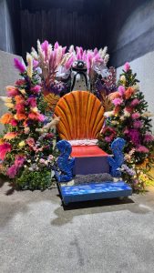 A whimsical display featuring a large, colorful shell-shaped throne in the center, adorned with vibrant pink, purple, and orange flowers and foliage. 