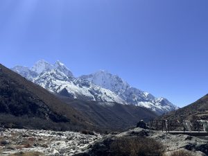 A panoramic view of snow-capped mountains under a clear blue sky, with rolling hills and rocky terrain in the foreground. 