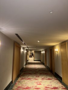 A long hotel corridor with beige walls and red patterned carpet. Soft lighting emanates from wall sconces along the sides, illuminating several wooden doors on both sides.