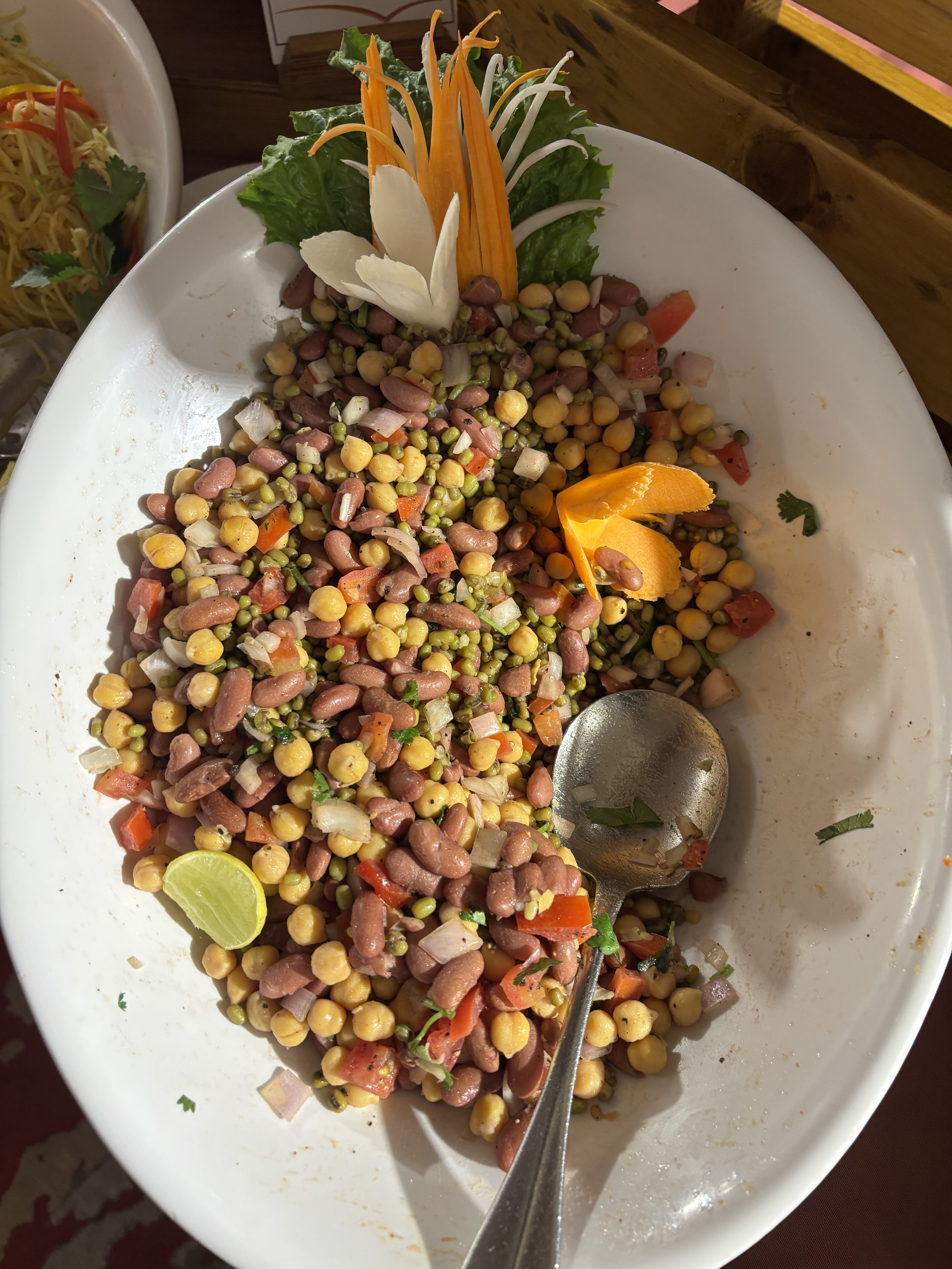 A white oval serving dish filled with a colorful salad made of mixed legumes, including yellow and red kidney beans, chickpeas, and green peas, combined with diced tomatoes, onions, and cilantro.