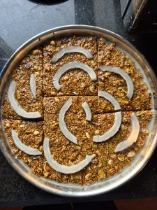 A round metal tray filled with square pieces of Chakku, a sesame seed and mixed nut dessert coated in glossy syrup.