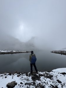 A person stands at the edge of Bhairav Kunda Lake, beside calm, mirror-like water, surrounded by a misty, fog-covered landscape in Sindhupalchok, Nepal. 
