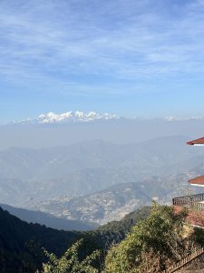 A panoramic view of snow-capped mountains against a blue sky filled with wispy clouds. Below, rolling hills and valleys stretch out, displaying a mixture of greenery and cultivated land. 
