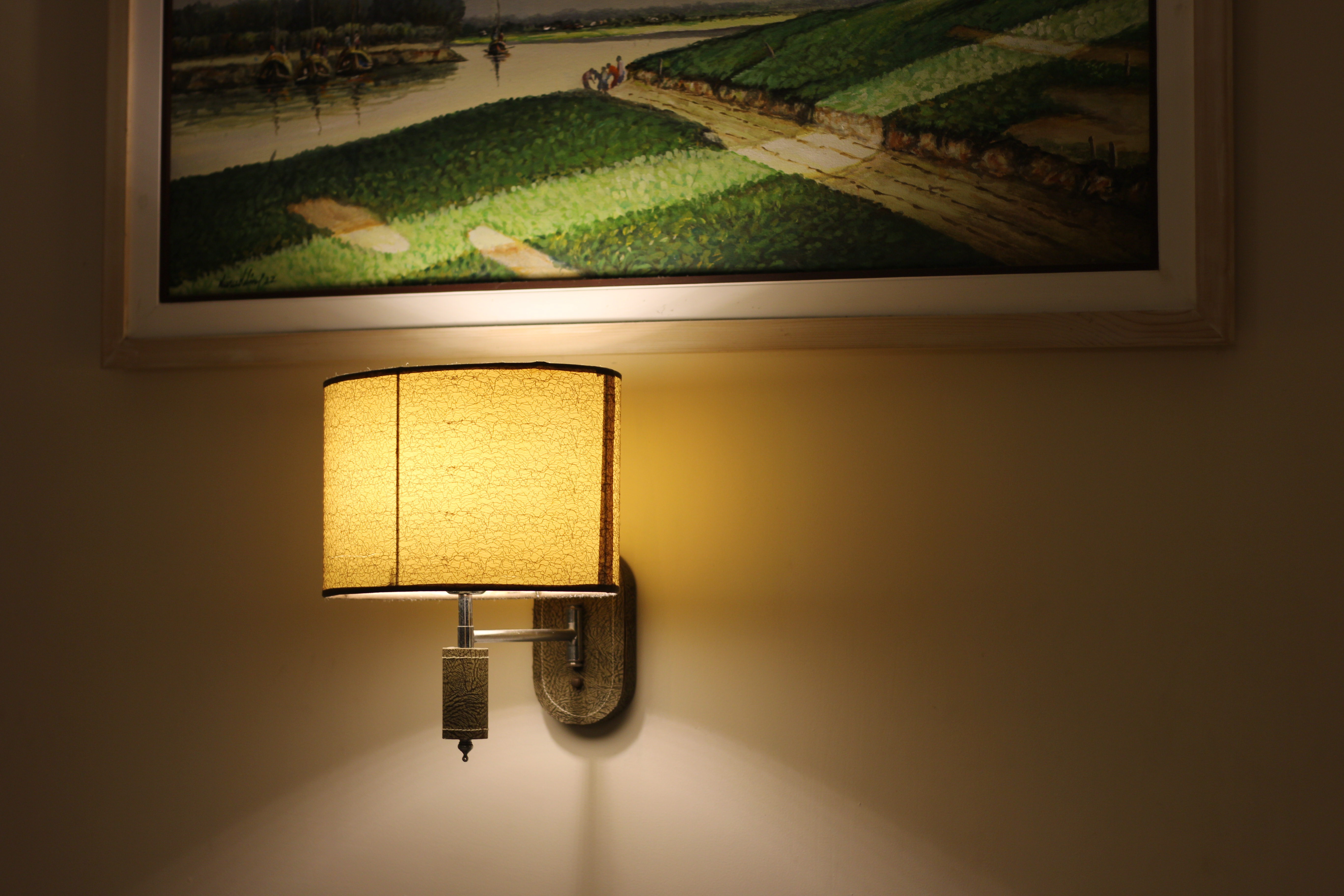 A decorative wall lamp with a beige shade emits a warm glow against a light-colored wall. Above the lamp, there is a framed painting featuring green fields and a river.
