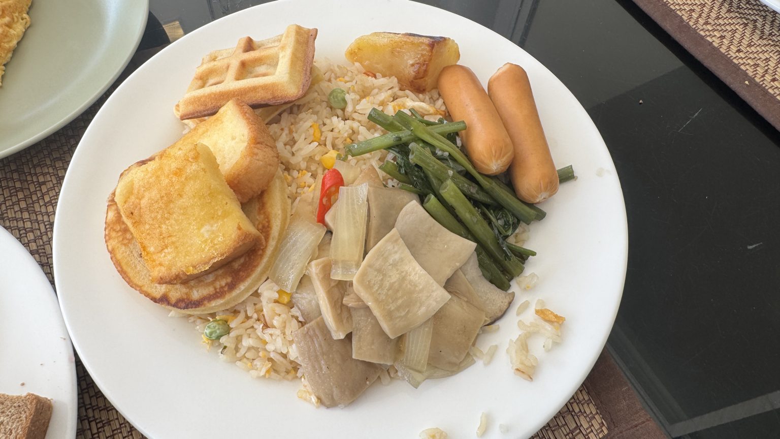A breakfast plate featuring fried rice, sausages, sautéed vegetables, and small waffles with pancakes.