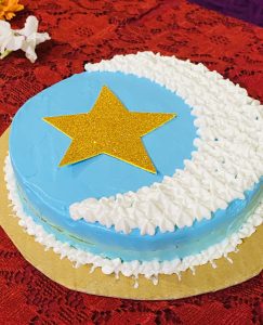 A round cake with light blue frosting features a textured white crescent design and a gold glitter star on top. It rests on a red lace tablecloth.