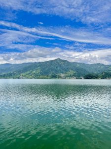 Clear blue water reflects green hills and a bright sky at a peaceful Phewa Lake in Pokhara, Nepal, showing calm weather and beautiful mountain scenery. 