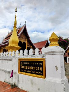 Entrance view of Vat Sensoukharam in Luang Prabang, Laos, showing a golden stupa, traditional red-roofed temple hall, and the monastery sign on a white boundary wall, reflecting classic Lao Buddhist architecture and sacred heritage.