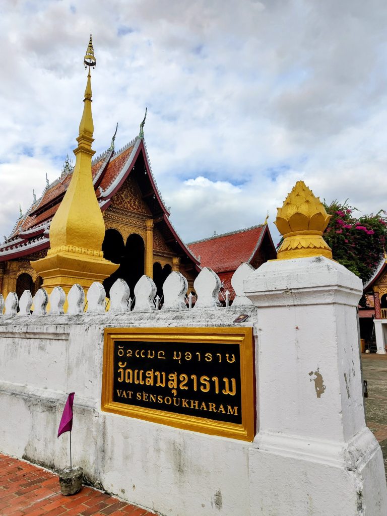 Entrance view of Vat Sensoukharam in Luang Prabang, Laos, showing a golden stupa, traditional red-roofed temple hall, and the monastery sign on a white boundary wall, reflecting classic Lao Buddhist architecture and sacred heritage.