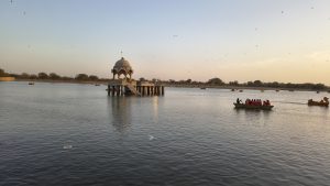 sandstone chhatri stands in the center of Gadisar Lake in Jaisalmer at sunset. Its domed architecture and a small staircase are reflected in the calm water. People in orange life jackets ride in rowboats and swan-shaped paddle boats nearby. The distant shore is lined with trees under a pale sky filled with a few birds.