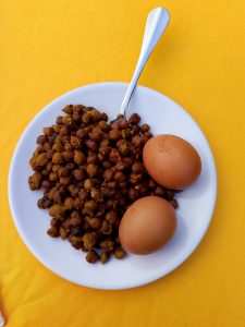 A white plate contains a serving of dark brown chickpeas, sprinkled with spices, alongside two whole, unpeeled brown eggs. 