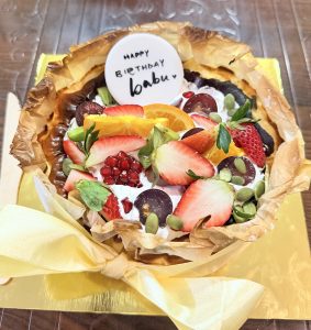 A round birthday cake decorated with a colorful assortment of fresh fruits, including strawberries, orange slices, pomegranate seeds, and grapes. 