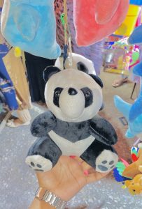 A hand holding a plush panda toy with a small smile, amidst colorful hanging toys. Bright and cheerful market setting, conveying a playful tone.