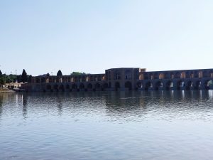 A calm spring view of Khaju Bridge in Isfahan, reflected in the Zayandeh Rud River. The historic brick arches stand under a clear blue sky, showing the beauty of Persian art and nature.