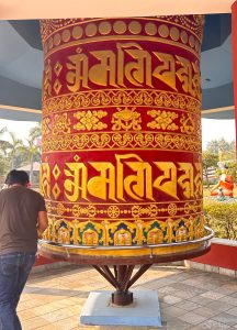 A large, colorful prayer wheel stands prominently, decorated in vibrant red and gold hues. 
