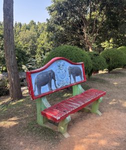 
A colorful bench in a park, featuring a blue backrest adorned with painted elephants on each side.