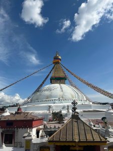 A large stupa adorned with colorful prayer flags, towering against a bright blue sky with a few fluffy clouds.