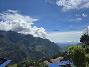 A panoramic view of a lush green mountainous landscape under a clear blue sky, dotted with fluffy white clouds. 
