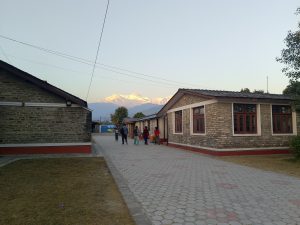 Stone houses line a paved walkway where people stroll along the path. Snow-capped mountains glow in the soft sunset light, creating a peaceful rural scene.