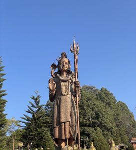 A large bronze statue of Lord Shiva stands against a clear blue sky, holding a trident with a serpent coiled around his neck.