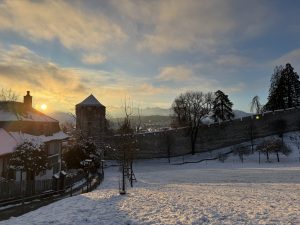 A winter sunrise over the snow-covered Museggmauer in Lucerne, Switzerland, featuring the Schirmerturm tower with the Alps in the background.