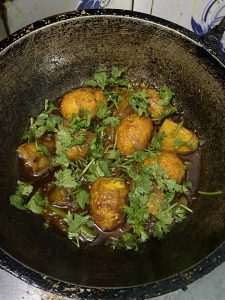 A savory dish in a black pan features golden-brown eggs in a rich brown gravy, garnished with fresh green coriander. The setting feels rustic and appetizing.