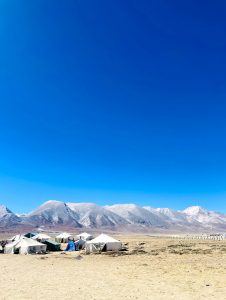 A wide, open landscape featuring several white tents in the foreground, set against a backdrop of majestic mountains under a clear blue sky. 