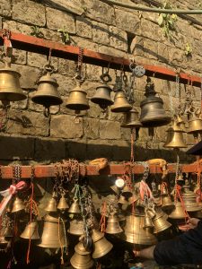 Two boards hanging horizontally in front of a stone brick wall.  From each board are hanging dozens of brass bells. There are also padlocks and some bananas left as offerings.
