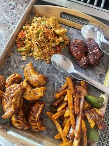 A variety of fried foods on a wooden tray.  In the top left is a stir fry with short curly noodles and vegetables. Top right are two small fried chicken wings with sesame seeds on them. Bottom left are fried dumplings with a light sauce and sesame seeds.  Bottom left are some things that look like french fries covered in a sauce and mixed with some green pepper slices. There are two forks and two spoons on the tray as well.