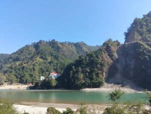 
A serene landscape featuring a river flowing alongside a rocky shoreline, surrounded by green hills and mountains. A white building with a red roof is visible on the left side, nestled among lush foliage.