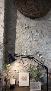 A cozy, creative workspace with a textured wall of notes and drawings, a circular woven piece, a desk lamp, plants, and two illustrated panels on a wooden desk.