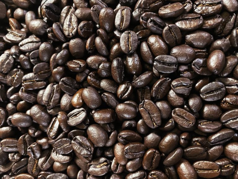 A close-up view of a pile of dark roasted coffee beans, showcasing their glossy, rich appearance and varying shapes, set against a dark backdrop.