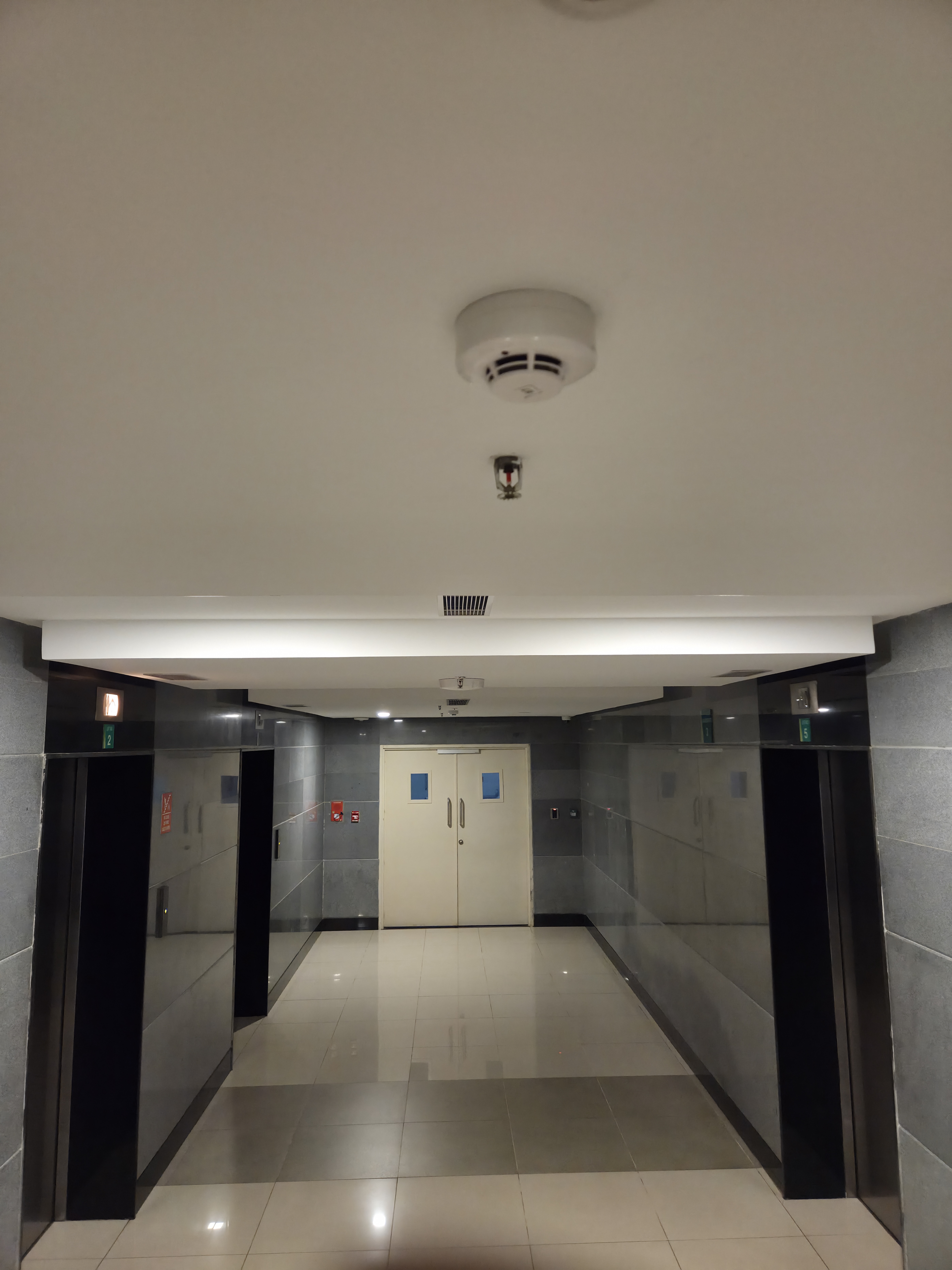 A hallway with glossy gray tiled walls, a white ceiling with a smoke detector, elevator doors on the left, and closed double doors with blue windows at the end.
