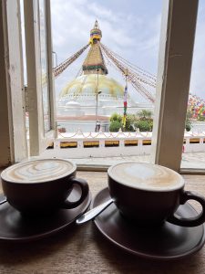 A view from an open window showcasing two cups of coffee with intricate latte art on wooden tables. In the background, the Boudhanath Stupa, a significant Buddhist monument, is visible, with its golden dome and prayer flags fluttering in the breeze under a partly cloudy sky.